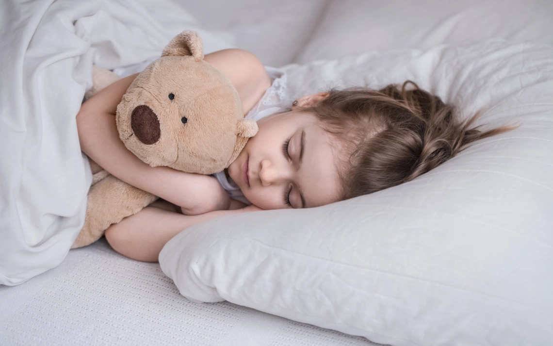 A young girl sleeps soundly in a comfortable bed with a teddy bear, illustrating peaceful rest.